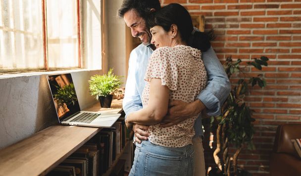 όμορφο ώριμο ζευγάρι αγκαλιάζει στέκεται ενώ κοιτάζοντας φορητό υπολογιστή στο σπίτι - έννοια της κλήσης βίντεο, online ψώνια - - Φωτογραφία, εικόνα