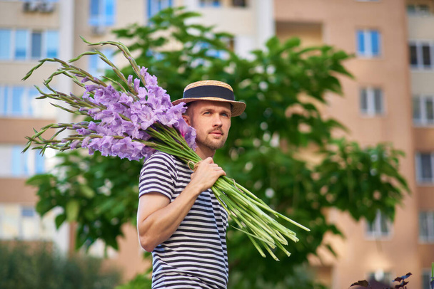мужчина улыбается, держа букет фиолетовых цветов гладиолусов, смотрящих в камеру на фоне зеленого сада. летняя концепция Высокое качество фото - Фото, изображение