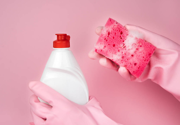 Ręce kobiety w różowych gumowych rękawiczkach trzymają mydlaną gąbkę kuchenną i detergent na różowym tle. Koncepcja zmywania naczyń. Zbliżenie. Skupienie selektywne. - Zdjęcie, obraz