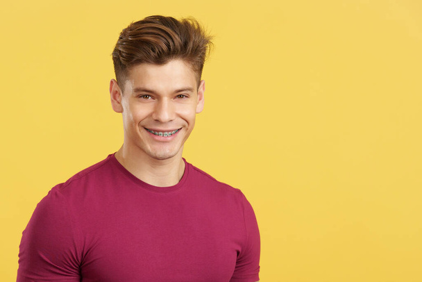 Caucásico hombre sonriendo mientras llevaba soportes en los dientes en el estudio con fondo amarillo - Foto, imagen