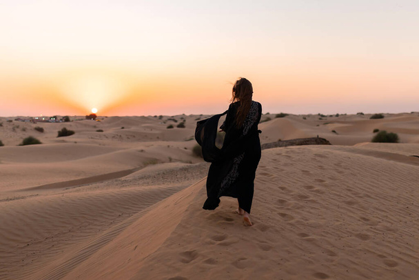 Beautiful mysterious woman in traditional arabic black long dress stands in the desert on sunset - Photo, Image