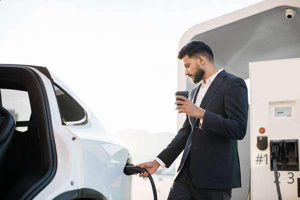 Side view of handsome arabian man in formal outfit holding coffee-to-go in one hand while inserting plug into luxury electric car. Charging process of modern eco transport. - Foto, Imagem