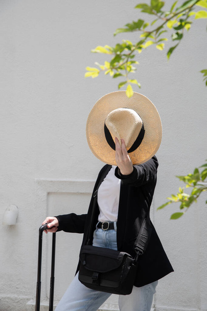 young happy brunette girl in sunglasses and straw hat with a suitcase goes on vacation, a woman stands against the background of a white brick wall, High quality photo - Foto, Imagen