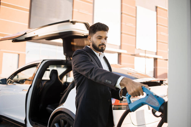 Good-looking indian man in business wear standing at charging station and taking electrical connector for charging battery of modern car. Alternative transport and ecology concept. - Zdjęcie, obraz
