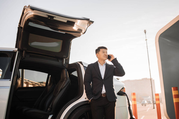 Успешный азиатский бизнесмен ведет мобильный разговор, ожидая, пока электромобиль подзарядит аккумулятор. Зрелый мужчина в стильном костюме опирается на роскошный белый автомобиль, смотрит в сторону и разговаривает по мобильному телефону. - Фото, изображение