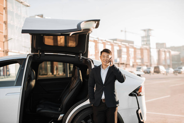 Successful asian businessman having mobile conversation while waiting for electric car to recharge battery. Mature man in stylish suit leaning on luxury white car, looking aside and talking on mobile. - Фото, изображение