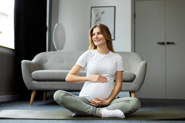 Ritratto di donna incinta sorridente seduta sul pavimento e che si tocca la pancia. Concetto di gravidanza yoga - Foto, immagini