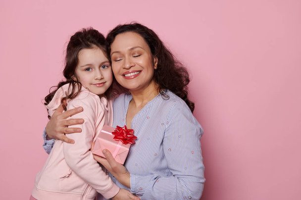 Feliz encantadora mujer multiétnica sonriendo una sonrisa dentada, gentil abrazando a su hermosa niña, posando con los ojos cerrados con un lindo regalo para el Día de las Madres, sobre un fondo rosa aislado - Foto, imagen