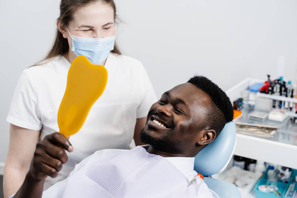 Afrikai férfi nézi a fogakat a tükörben a fogorvosnál. Boldog afro-amerikai fogorvosi beteget. Fogkezelés. Jóképű férfi ül a fogorvosi rendelőben és mosolyog. - Fotó, kép
