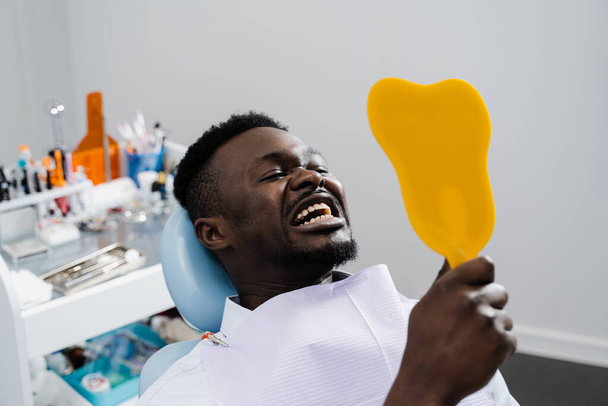 Consulta con dentista en clínica dental. Un paciente afroamericano está visitando al dentista. Hombre africano paciente está mirando en el espejo en sus dientes después de quitar la caries y llenar los dientes - Foto, Imagen