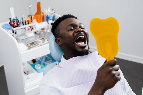 Konsultacja z dentystą w klinice stomatologicznej. Amerykański pacjent odwiedza dentystę. Pacjent afrykański patrzy w lustro po usunięciu próchnicy i wypełnieniu zębów. - Zdjęcie, obraz