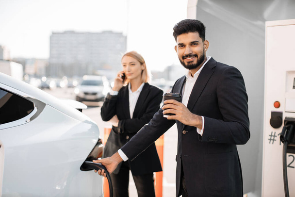 Happy caucasian business lady having phone call outdoors while her indian male partner charging white electric car. Business trip on luxury hybrid auto with zero emission. - Foto, Bild