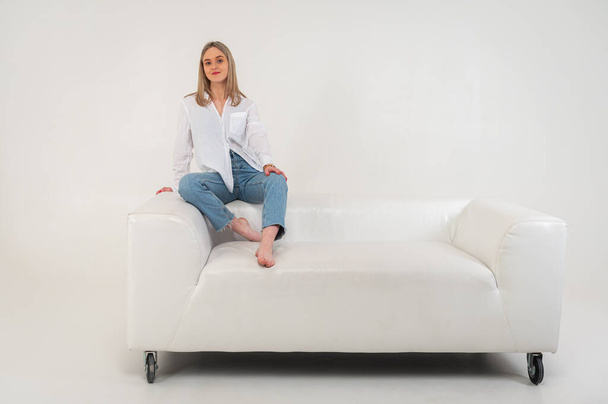 Happy young girl relaxes on comfortable soft white leather sofa, calm contented girl stretches on sofa, thinking about pleasant things. Isolated on light background, place for inscription - Zdjęcie, obraz
