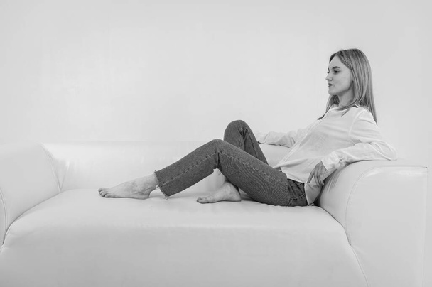 Чорно-біла класика, молода дівчина розслабляється на зручному м'якому білому шкіряному дивані, спокійна задоволена дівчина розтягується на дивані. Ізольовано на фоні світла, місце для тексту
 - Фото, зображення