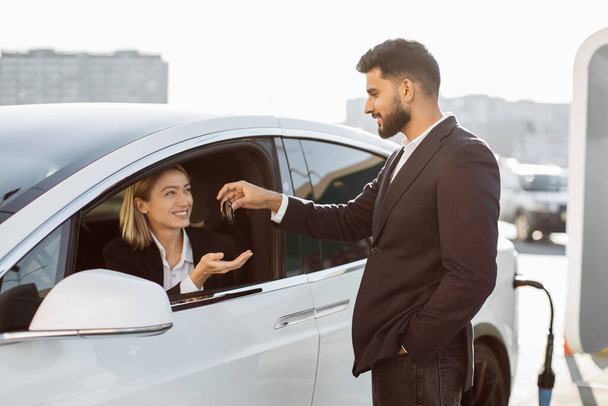 Cheerful caucasian woman sitting in new modern car while indian seller standing near and giving her keys. Business lady in stylish suit buying vehicle from professional consultant. - Photo, image