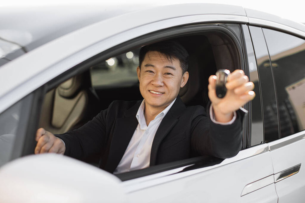 Satisfied asian man in business suit showing car keys while sitting inside. Handsome successful businessman buying new transport at modern dealership. - 写真・画像