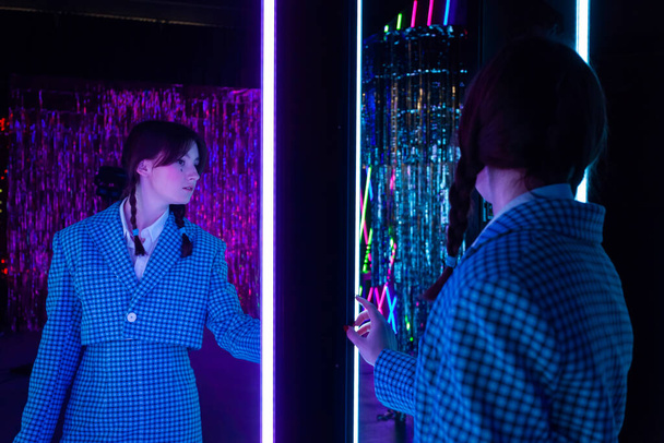 A cute girl with drawn tears, dressed in a blue plaid jacket and skirt with knee high socks, looks mysteriously at herself in the mirror in a neon-lit room. - Foto, afbeelding