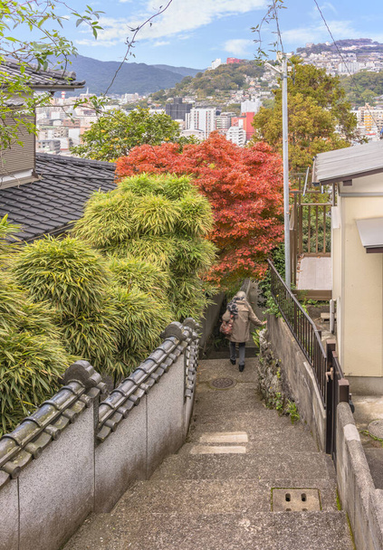 Starsza kobieta schodząca w dół stromego zbocza z wąskim schodem schodzącym wzgórzami Nagasaki między Japońskimi domami których ogrodzenie pozwala bambusowym krzewom i czerwonym momiji klonowym drzewom wystawać jesienią - Zdjęcie, obraz