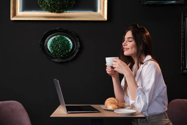 Profil einer jungen Unternehmerin, die in einem Café sitzt und ihren Morgenkaffee genießt. Es gibt ein Frühstück auf einem Tisch und einen Laptop. - Foto, Bild