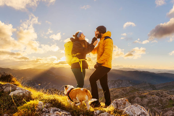 пара туристів з рюкзаками і собака, що розглядає пейзаж після досягнення вершини гірського піку. чоловік і жінка гуляють на заході сонця. спорт на відкритому повітрі
. - Фото, зображення