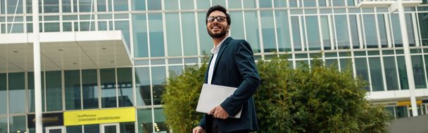 Uomo d'affari sorridente in giacca e cravatta con computer portatile vicino a un moderno edificio per uffici sta guardando lato - Foto, immagini
