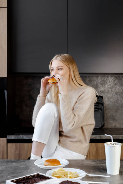Een mooi slank jong blond meisje zit in een moderne keuken en eet een hamburger, fastfood en snoep. contrast tussen een ongezond dieet en een schoonheid. - Foto, afbeelding