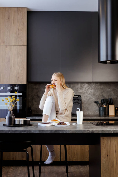 A beautiful slender young blonde girl is sitting in a modern kitchen and eating a burger, fast food and sweets. contrast between an unhealthy diet and a beauty. - Foto, Imagen