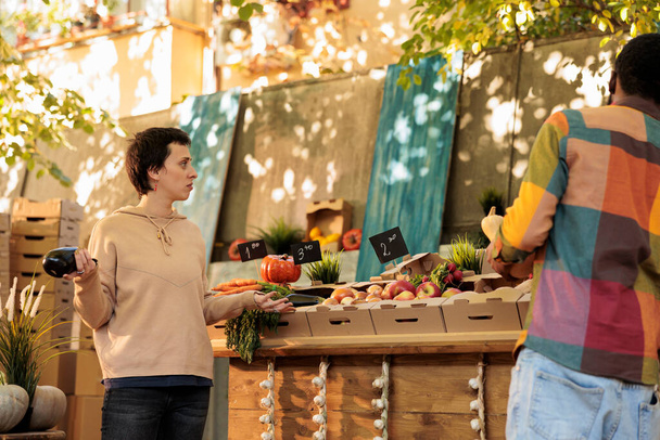 Молодая семья разговаривает друг с другом и покупает здоровые органические овощи на фермерском рынке, местном продовольственном киоске. Люди покупают свежие натуральные продукты, смотрят на органические продукты. - Фото, изображение