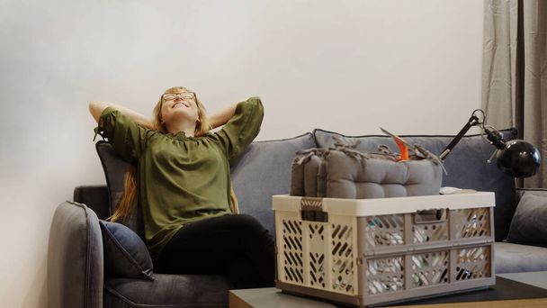 happy woman resting on the couch after moving in and a box on the table, satisfied homeowner enjoying her first day in new house. High quality photo - Photo, Image
