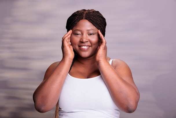 African american body positive model touching fresh healthy skin and looking at camera with happy facial expression. Beautiful plus size woman posing in studio for skincare cosmetology - Foto, imagen