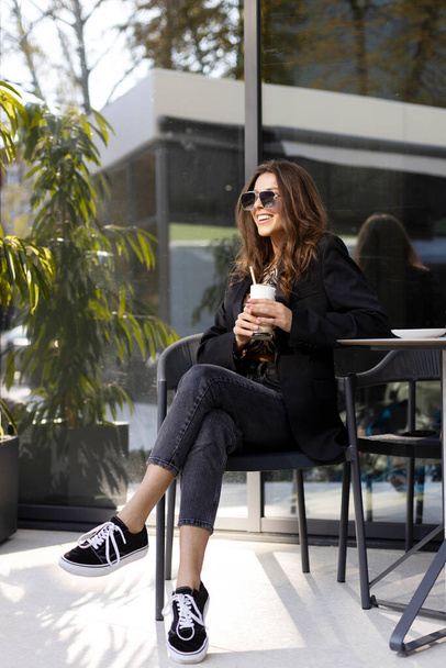 Positief, lachend meisje met een zonnebril zit aan een tafel op straat, in een café, gekruiste benen. Zelfverzekerde zakenvrouw die koffie drinkt, concept koffiepauze, lunchpauze, rust. - Foto, afbeelding
