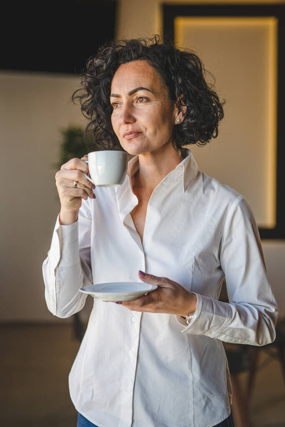 Yksi nainen seisoo ikkunan kotona tai toimistossa liikenainen yrittäjä käyttää valkoista paitaa ottaa kuppi kahvia vakava huolissaan oikeita ihmisiä kopioida tilaa aamulla rutiini kypsä valkoihoinen nainen - Valokuva, kuva