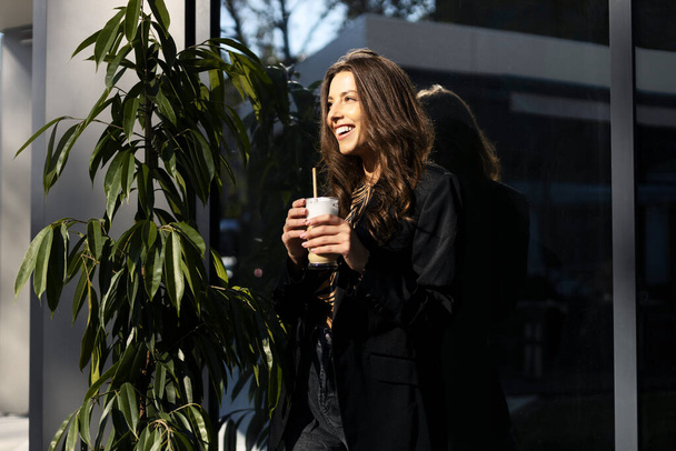 Sorridente ragazza bruna beve latte o cappuccino, tiene una tazza in mano. Attraente giovane donna rilassante in un caffè. - Foto, immagini