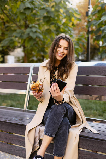 Beautiful girl is sitting on a bench, holding a mobile phone, taking a selfie. A young girl is holding a croissant in her hand, making a video for her followers. Girl influencer. - Foto, Bild