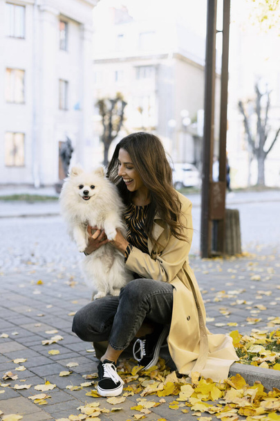 Привлекательная молодая девушка держит красивую собаку на руках, на улице. Улыбающаяся девушка держит своего питомца, опавшие листья, осень. - Фото, изображение