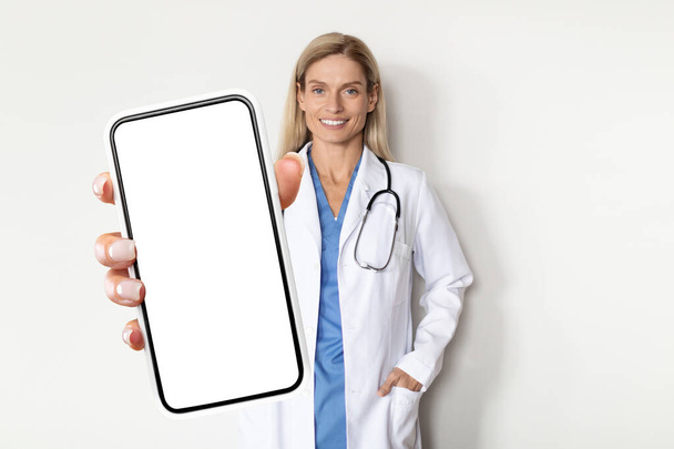 Mujer sonriente del doctor en uniforme que demuestra la pantalla grande del teléfono inteligente en blanco en la cámara mientras que está parado cerca de la pared blanca en el hospital, médico femenino que muestra el espacio de copia para el anuncio médico, burla - Foto, imagen