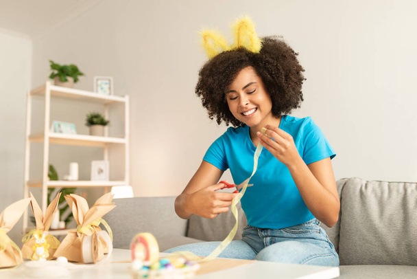 Hermosa joven hembra negra haciendo decoraciones de Pascua en casa, mujer afroamericana feliz usando diadema de orejas de conejo preparando regalos festivos Embalaje, corte de cinta y sonrisa, espacio libre - Foto, imagen