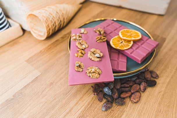 Various ruby pink chocolate bars with nuts, dried orange slices and toasted cacao beans on plate. Wooden table. High quality photo - Foto, immagini