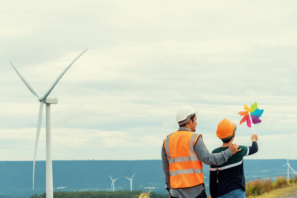 Engineer with his son holding windmill toy on a wind farm atop a hill or mountain. Progressive ideal for the future production of renewable, sustainable energy. Energy generated from wind turbine. - Photo, image