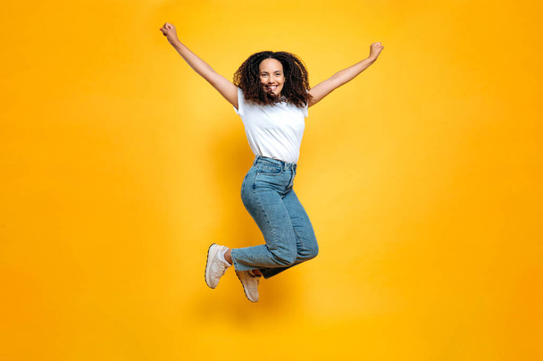 Full length photo of cheerful mixed race young woman with curly hair dressed in casual clothes, bobbing high on isolated orange background, looking at camera, smiling happily - Photo, Image
