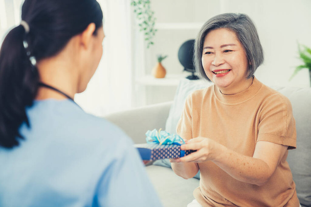 A young caregiver hand over to her senior patient a blue gift box with blue ribbons at a contented living room. - Fotoğraf, Görsel