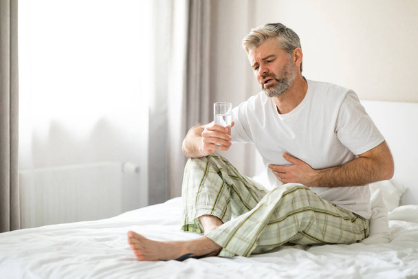 Unhappy sick middle aged man sitting on bed with glass of water and touching his belly, suffering from stomach pain in the morning, wearing pajamas, home interior. Gastrointestinal Diseases, Problems - Zdjęcie, obraz