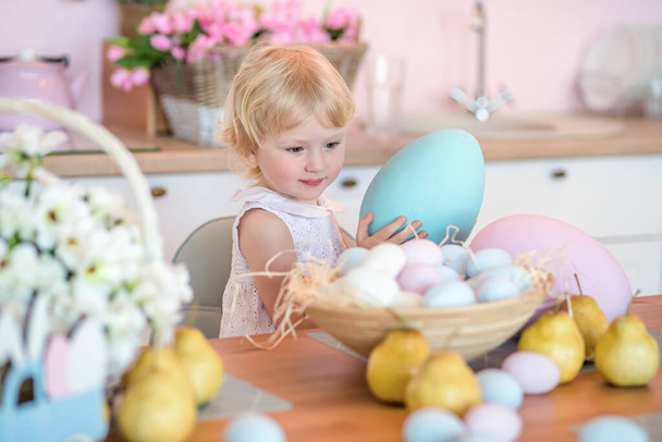 Une petite fille sort un gros œuf de Pâques peint dans un panier rempli d'œufs de mcolorfu. Un enfant dans une cuisine décorée pour Pâques après la peinture - Photo, image
