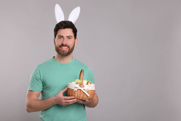 Portrait of happy man in cute bunny ears headband holding wicker basket with Easter eggs on light grey background. Space for text - Foto, Bild