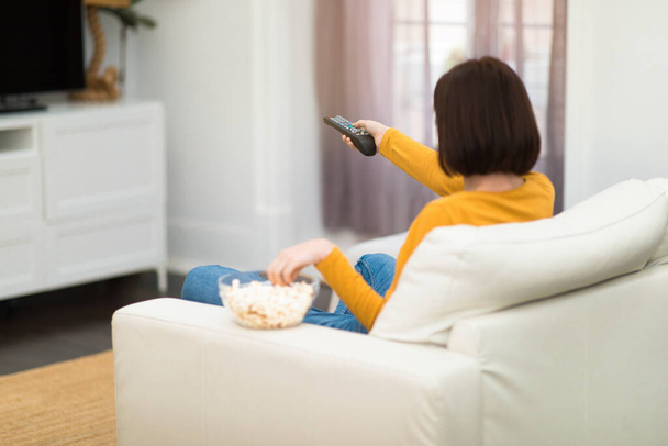Πλάτη άποψη της μελαχρινής νεαρής γυναίκας σε άνετο casual outwear κάθεται στον καναπέ στο σαλόνι, κρατώντας τηλεχειριστήριο, τρώγοντας ποπ κορν, αλλάζοντας κανάλια στην τηλεόραση, περνώντας το Σαββατοκύριακο στο σπίτι, αντίγραφο χώρου - Φωτογραφία, εικόνα