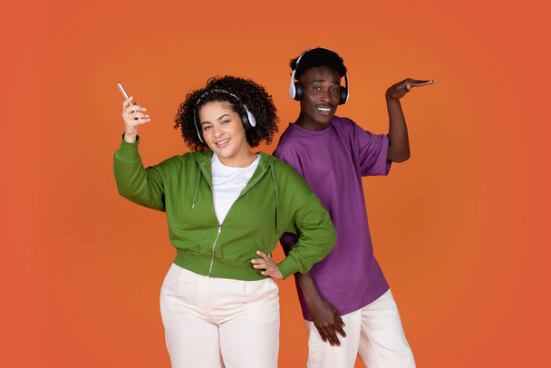 Joyful funny happy young multiethnic couple african american guy and hispanic lady listening to music and dancing together on red background, using smartphone and wireless headphones - Valokuva, kuva