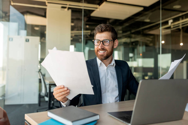 Happy male entrepreneur reading business papers and smiling, positive businessman holding documents, checking company growth rates, sitting at desk in office - Photo, Image