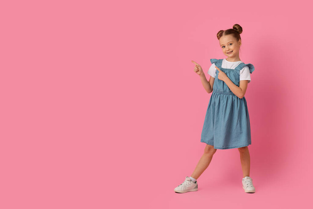 Spójrz na to. Cute Little Girl wskazując obok na miejscu skopiować z dwoma palcami, stojąc nad różowym tle studio, uśmiechnięta kobieta dziecko demonstrujące wolne miejsce do projektowania lub oferty - Zdjęcie, obraz
