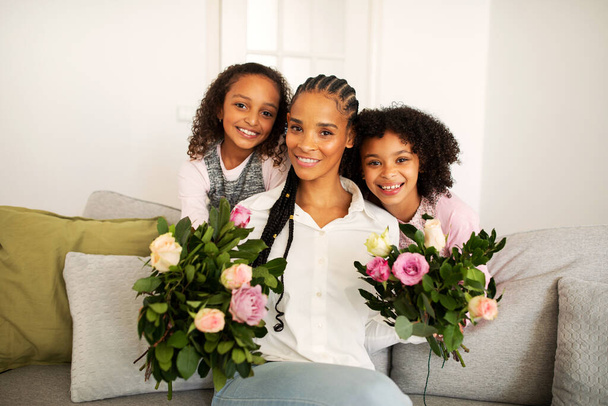 Glückliche schwarze Mutter mit Blumen, die Glückwünsche von ihren hübschen Töchtern entgegennimmt, die zu Hause auf der Couch sitzen und in die Kamera lächeln. Konzept für Familienfeier - Foto, Bild