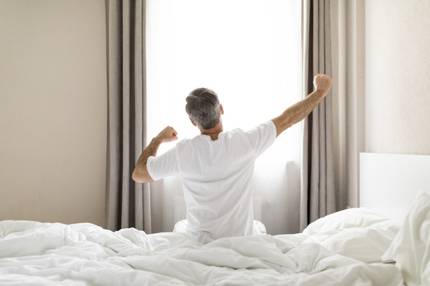 Rear view of unrecognizable middle aged man in pajamas sitting on bed and stretching body after waking up in the morning, looking at window, copy space. Comfortable healthy sleep concept - 写真・画像